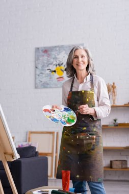 positive middle aged woman holding paintbrush and palette with colorful paints near easel  clipart