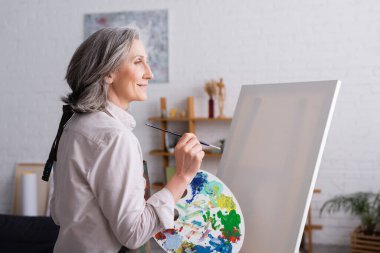 Orta yaşlı bir kadın elinde boya fırçası ve palet ile boş tuvalin yanında renkli boyalar tutuyor. 