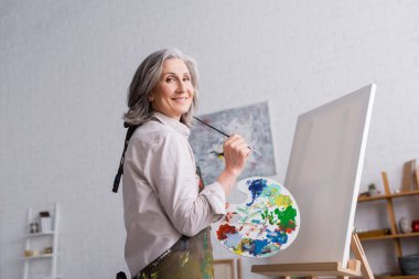 neşeli orta yaşlı kadın elinde boya fırçası ve palet ile boş tuvalin yanında renkli boyalar tutuyor.