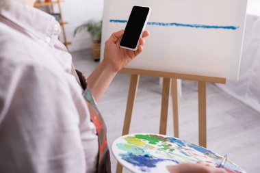 Olgun bir kadının palet, boya fırçası ve arka planda boş ekranlı akıllı telefonu tutuşu kısmen görülüyor. 