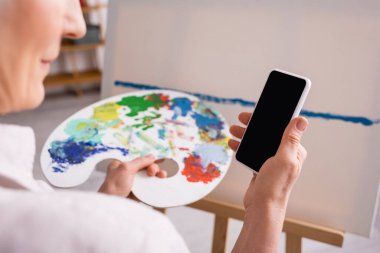 Esmer, olgun bir kadının palet ve akıllı telefon tutarken görüntüsü bulanık arkaplandaki boş ekranın yanında. 