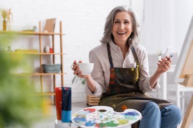 Orta yaşlı mutlu ressam elinde boya fırçası, bir fincan kahve ve tuvalin yanında akıllı telefon kullanıyor. 