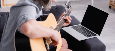 Bulanık orta yaşlı bir kadının laptopun yanında akustik gitar çalmayı öğrenmesi. Kanepede boş ekran, pankart.