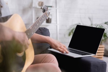 Bulanık orta yaşlı kadının akustik gitar çalmayı öğrenme görüntüsü. Kanepede boş ekran ile dizüstü bilgisayar.