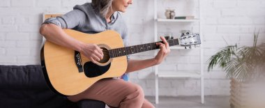 Oturma odasında otururken akustik gitar çalan gri saçlı olgun bir kadının kısmi görüntüsü, afiş.