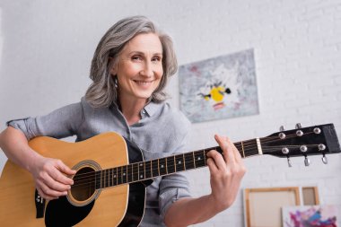 Gülümseyen orta yaşlı kadın oturma odasında akustik gitar çalıyor.