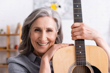 Oturma odasında elinde akustik gitar tutan gri saçlı mutlu olgun bir kadın.