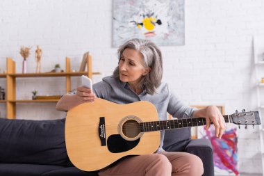 Akustik gitar çalmayı öğrenirken akıllı telefona bakan gri saçlı olgun bir kadın. 