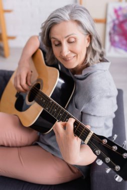 Oturma odasında akustik gitar çalan gri saçlı olgun bir kadın.