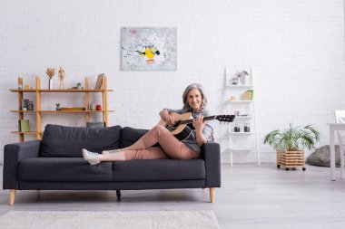 Mutlu, gri saçlı, koltukta oturan ve modern oturma odasında akustik gitar çalan olgun bir kadın.