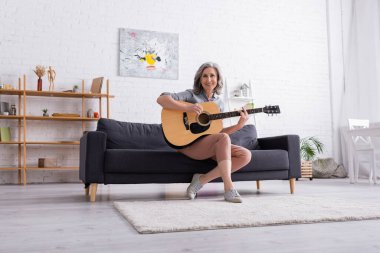 Kanepede oturan ve oturma odasında akustik gitar çalan gri saçlı neşeli olgun bir kadın.