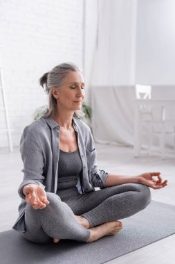 mature woman with grey hair sitting in lotus pose while meditating on yoga mat clipart