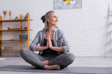 happy mature woman with grey hair sitting with praying hands in lotus pose on yoga mat clipart