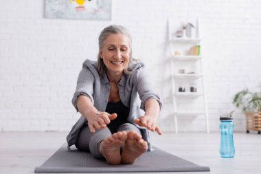 Mutlu, olgun, gri saçlı bir kadın yoga minderinin üzerinde spor şişesinin yanında geriniyor. 