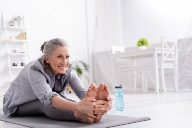 Neşeli olgun kadın, gri saçlı yoga minderinin üzerinde spor şişesinin yanında geriniyor. 