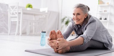 Spor şişesinin yanında yoga minderinde gerinen gri saçlı mutlu olgun kadın, afiş.