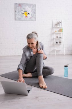Yoga minderinde oturmuş laptopta özel ders izleyen gri saçlı olgun bir kadın.