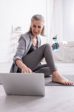Gri saçlı olgun bir kadın yoga minderinde oturmuş dizüstü bilgisayar izliyor ve elinde spor şişesi tutuyordu.