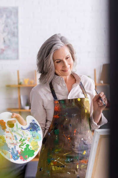cheerful mature woman in apron holding paintbrush and palette while looking at canvas