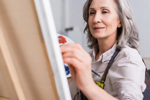 Mature Woman Holding Paintbrush While Painting Canvas Blurred Foreground — Stock Photo, Image