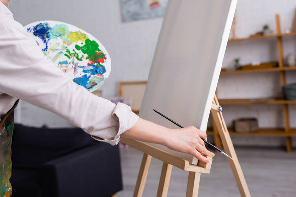 partial view of middle aged woman holding paintbrush and palette with colorful paints near blank canvas 