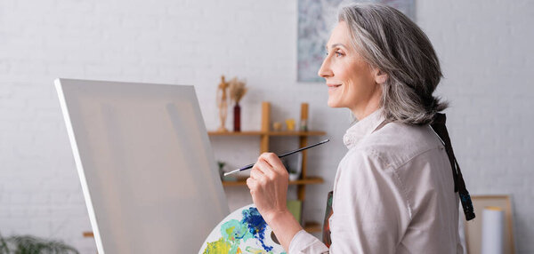 довольная женщина средних лет, держащая кисть и палитру с красочными красками рядом с чистым холстом, баннер