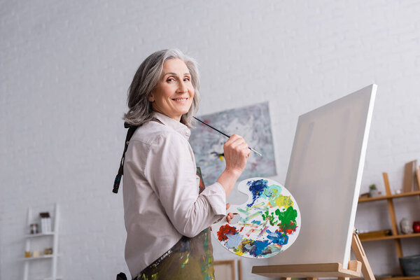 cheerful middle aged woman holding paintbrush and palette with colorful paints near blank canvas