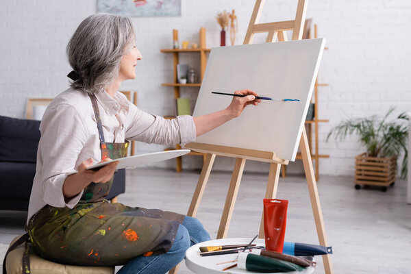 cheerful middle aged woman holding paintbrush and palette while painting on canvas
