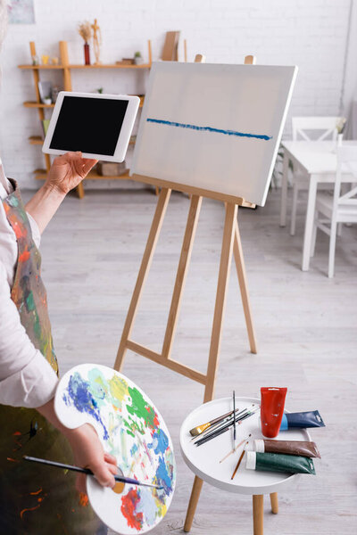 cropped view of mature woman holding palette and digital tablet with blank screen near canvas