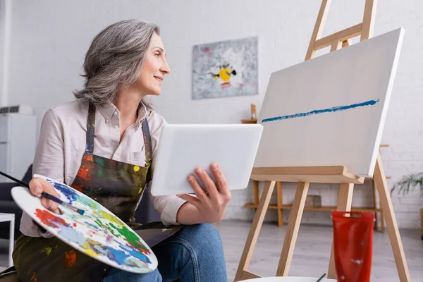 Pleased Mature Woman Holding Palette Digital Tablet While Looking Canvas — Stock Photo, Image