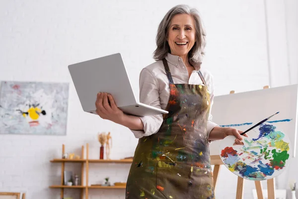 Positive Mature Artist Holding Palette Laptop While Watching Tutorial Canvas — Stock Photo, Image