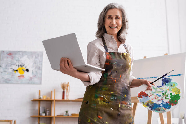 положительный зрелый художник держит палитру и ноутбук во время просмотра учебника возле холста