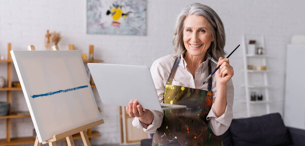 счастливый зрелый художник, держа кисть и ноутбук рядом с канделябрами, баннер