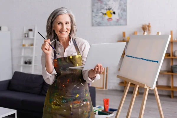 Happy Mature Artist Holding Paintbrush Laptop Canvas — Stock Photo, Image