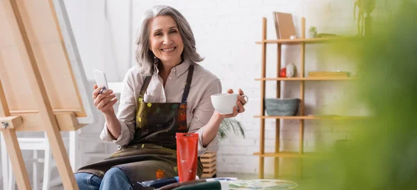 Happy Middle Aged Artist Holding Paintbrush Cup Coffee Using Smartphone — Stock Photo, Image