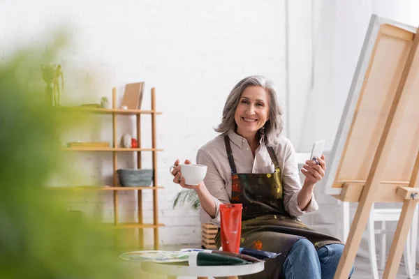 Happy Middle Aged Artist Holding Paintbrush Cup Coffee Using Smartphone — Stock Photo, Image