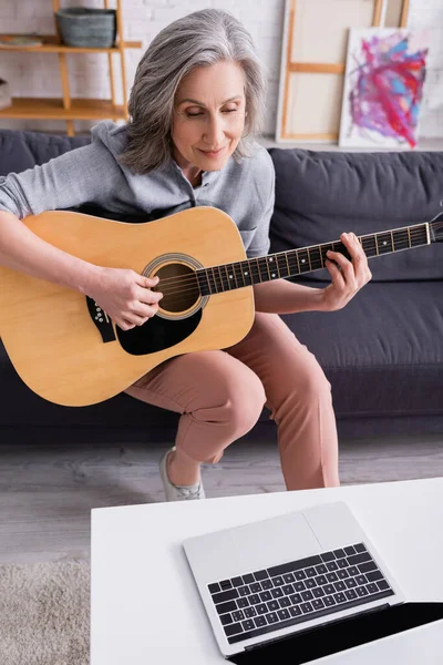 middle aged woman with grey hair learning to play acoustic guitar near laptop with blank screen on coffee table