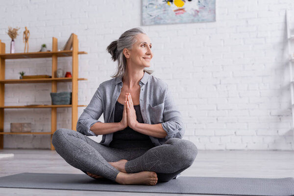 счастливая зрелая женщина с седыми волосами сидит с молящимися руками в позе лотоса на коврике для йоги