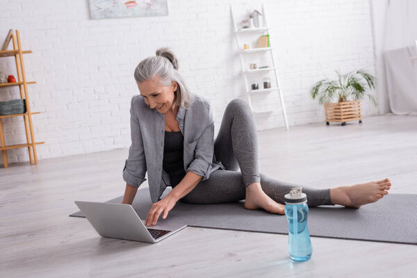 smiling mature woman sitting on yoga mat while watching tutorial on laptop 