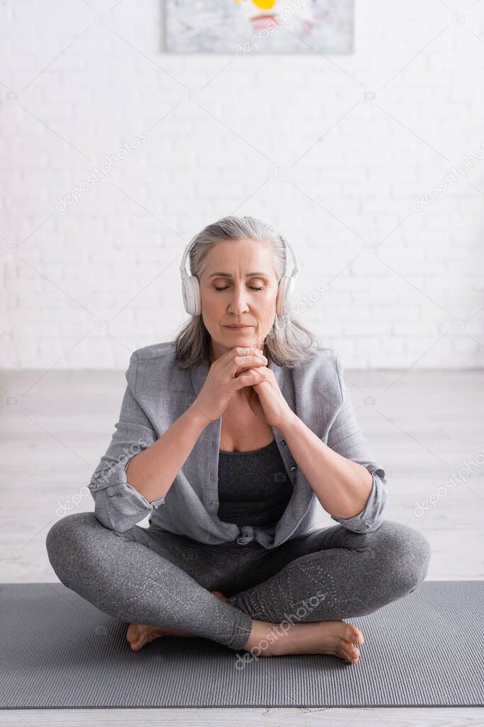 middle aged woman in wireless headphones meditating while sitting in lotus pose on yoga mat 