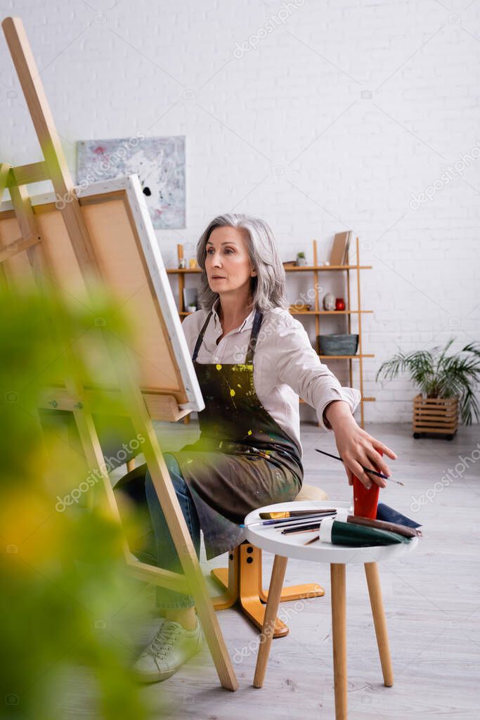 mature artist holding paintbrush and reaching paint in tube while sitting near canvas 