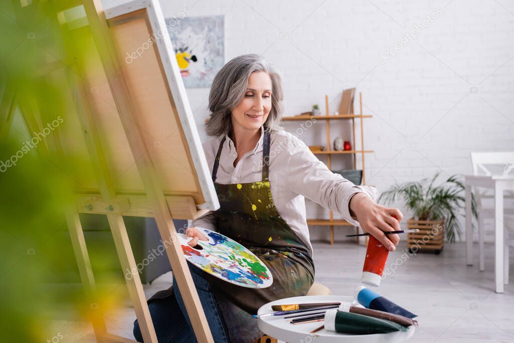 mature woman holding paintbrush and palette while reaching red paint in tube and sitting near canvas 