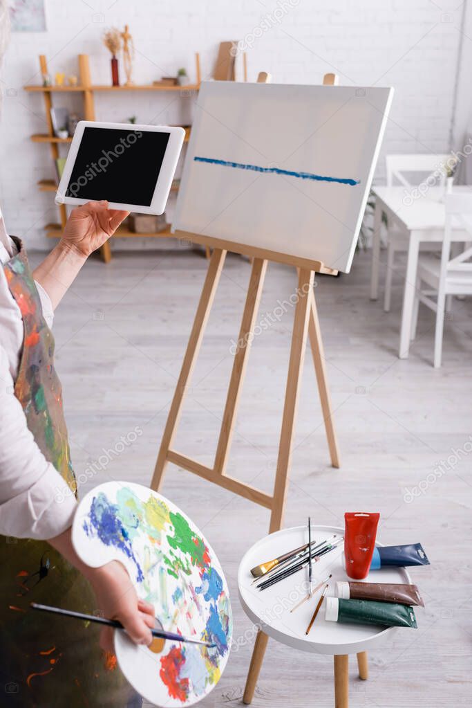 cropped view of mature woman holding palette and digital tablet with blank screen near canvas