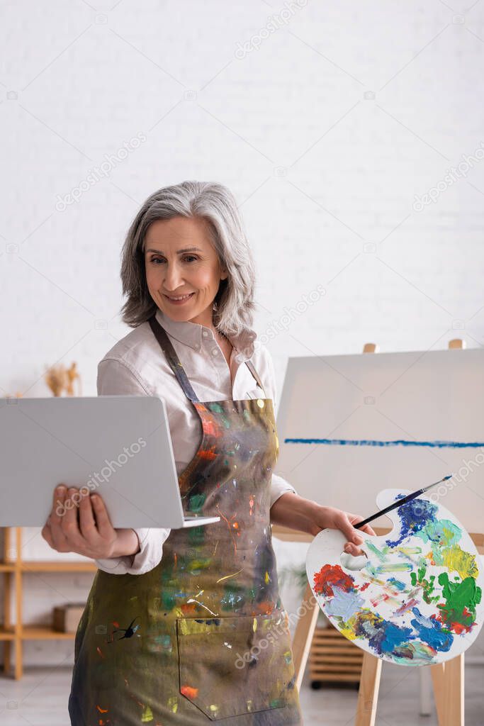 smiling mature woman holding palette and laptop while watching tutorial near canvas
