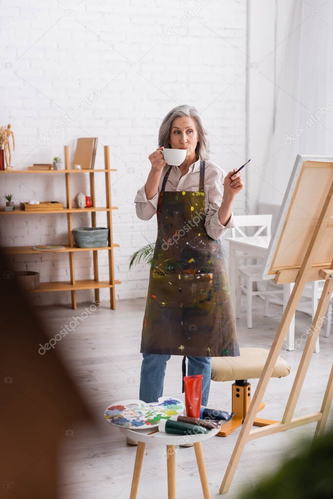 middle aged artist holding cup of coffee and paintbrush while looking at canvas 
