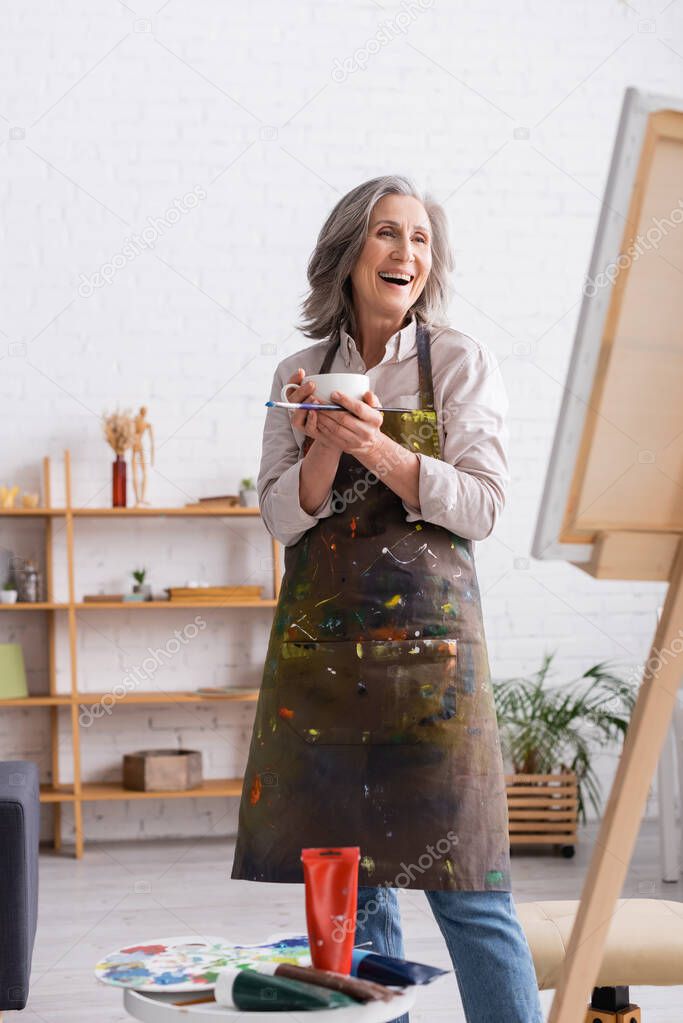 excited middle aged artist holding cup of coffee and paintbrush near canvas 