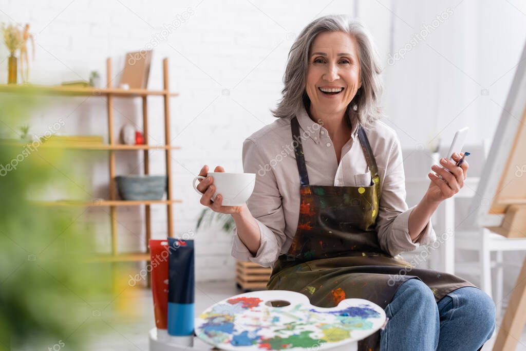 happy middle aged artist holding paintbrush, cup of coffee and using smartphone near canvas 