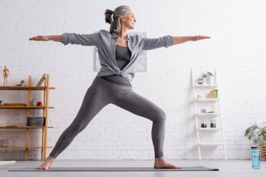Orta yaşlı spor elbiseli kadın evde yoga yaparken savaşçı pozu veriyor. 