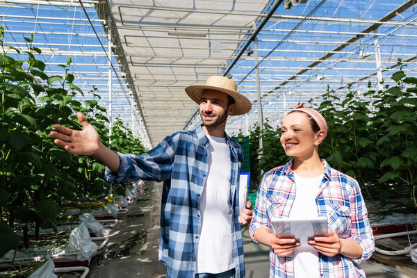 happy african american farmer holding digital tablet near colleague pointing with hand in greenhouse