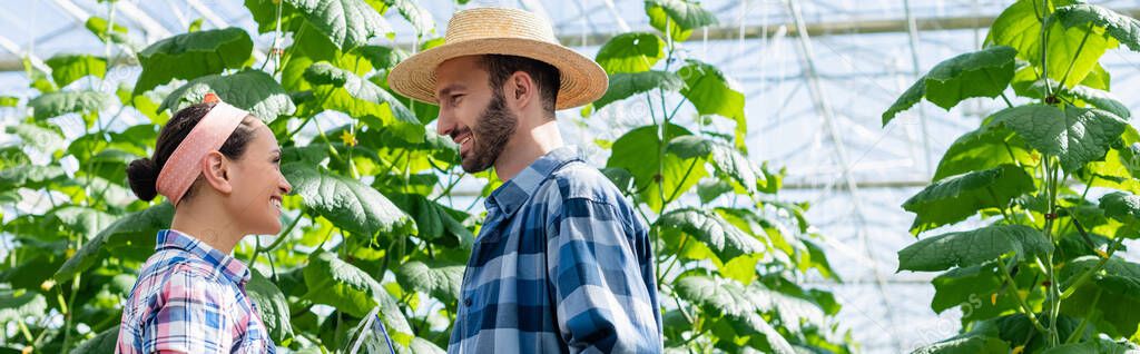 side view of smiling interracial farmers talking in greenhouse, banner